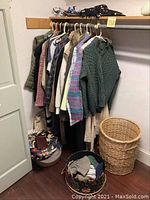 Photo shows a wooden clothing rack with multiple women's sweaters and coats hanging, a basket with clothes, a wicker basket, and handbags on the shelf above.