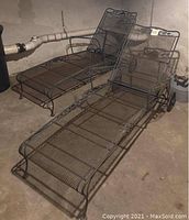 Two black wrought iron lounge chairs with mesh frame and wheels positioned side by side in a basement space with concrete floor and walls.