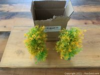 Two artificial aquarium plants with green and yellow leaves on teal bases placed in front of a cardboard box.