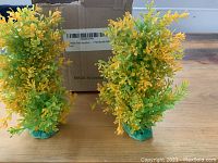 Two yellow artificial aquarium plants with green bases shown side by side on wooden surface in front of a cardboard box.