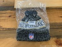 New black, white, and gold New Orleans NFL knit toque cap in plastic packaging, featuring NFL shield logo patch and 'New Orleans' knitted text, with removable pompom on top.