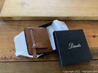 Brown textured leather wallet partially open displaying card slots and zippered compartment alongside black Dante branded box on wooden surface.