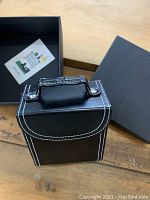 Black travel size jewelry case with white stitching and small handle next to outer box on wood surface.
