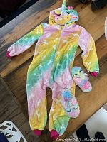 Full view of the rainbow unicorn pajama suit laid flat, with slippers placed beside it on wooden surface.