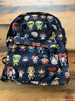 Front view of the backpack showing multiple cartoon Stranger Things characters, front zipper pocket, and logo patch.