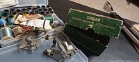 Wide view of plastic case with various thread spools, bobbins, and metal sewing attachments next to original Singer blind stitch hemmer box.