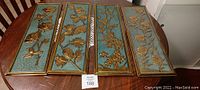 Four rectangular copper wall art panels arranged on a table. Each panel has embossed botanical designs and gold-colored frames on a textured blue-green background.