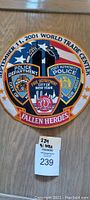 Photo of a large circular embroidered patch about 12 inches diameter, hung on a vertical surface. The patch commemorates fallen heroes of September 11, 2001 World Trade Center attacks, depicting insignias for Police Department, Port Authority Police, Fire Department of NYC, and mentions Hudson Valley and New Jersey.