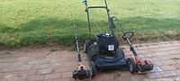 Overview of the Precision push lawn mower positioned in the center with two Homelife weed eaters on either side on a brick patio background