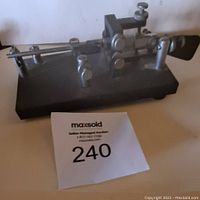 Side view of vintage telegraph key showing horizontal metal lever mechanism with adjustment screws on a black base with auction lot number 240.