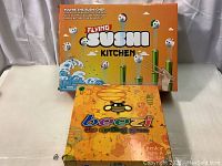 Photo showing both board games: Flying Sushi Kitchen in bigger box at back, Beezi The Spelling Game in smaller box front.