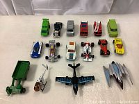 Top-down view of all 16 vehicles arranged in rows on a white surface, including cars, trucks, racing cars, and aircraft.