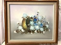 Framed oil painting of flower bouquet with white and blue blossoms spilling from a golden basket on a pastel background, signed by Ray.