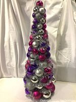 Full view of a 30-inch tall cone-shaped Christmas tree decorated densely with shiny round ornaments in silver, purple, and magenta.