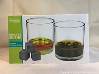 Front of product box showing two whiskey glasses with whiskey inside and three whiskey soap stone rocks in front.