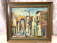 Framed oil painting showing a village street scene with buildings, archway, and lamppost, with two figures walking down the street.