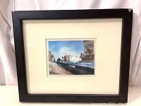 Black framed watercolor painting behind glass, showing full view with white matting and urban street scene.