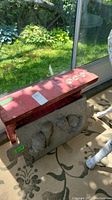 Side view of red solid wood bench with aluminum cat mold base, outdoors with garden background.