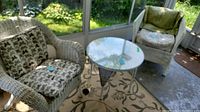 Two white wicker chairs with patterned cushions and a round white wicker table with glass top on enclosed porch.