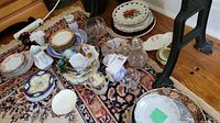 Photo displaying various plates, bowls, and kitchenware items arranged on a rug, including floral patterned plates and clear glass bowls.