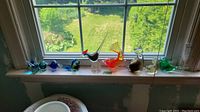 Windowsill display of eight glass figurines in a row: four small bird paperweights, two rooster figures, one cat and one large bird