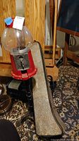 Photo of vintage red gumball machine with large clear glass globe, visible metal coin mechanism, and part of black metal stand. Trombone case visible in background.