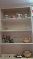 Photo of shelf with red and white pitcher, jug, two small matching cups, beige bowls, glass cookie jar, glass cake stand, box grater, cherry pitter, and small plates.