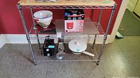 Wide view of the chrome wire shelving unit with wooden top showing all items on shelves including ceramic colanders, boxed toaster, frying pan, and Perfect Cooker appliance box.
