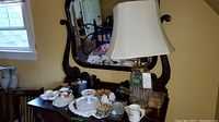 Wide shot showing the collection of items on a wooden dresser with mirror: dishes, camera, glass lamp, mugs.