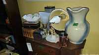 Photo showing desk lamp, wall mount lamp, ceramic jug, white decorative vase, copper mug, cane basket, tin box arranged on wooden drawer.