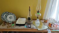 Wide view of all items on lace-covered table including three glass oil lamps, two collector plates on stands, brass candlestick holders, rectangular mirror tray, and colorful mugs.