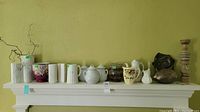 Shelf display with three white ceramic vases, a pink ceramic planter, teapots, milk jug, ceramic pitcher, glass vase, and metal art pieces.