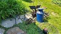 Wide outdoor view showing blue cast iron milk can, metal plant stands, round table with cast iron legs, log holder, antique wheel, and hand pump placed on patio with grass and fence background.