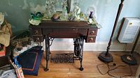 Full view of vintage New Williams treadle sewing machine table with cast iron legs, wooden top, and drawers. Decorations on top include glass figurines and small items.