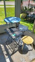 Full view of the vintage black metal table with mesh top and lower rack, black metal plant stand, and yellow wooden stool with knitted cushion.
