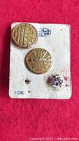 Three pins shown attached to the original 10K marked cardboard backing. The cardboard is worn and has some stains and surface damage. Two pins are gold, round with text and starburst design. One smaller pin is pentagon-shaped with blue and white enamel and gold star detail.