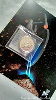 Silver coin in protective casing resting on Star Trek themed background with a spaceship image.