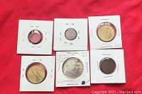 Six coins individually encapsulated in cardboard holders on red background including Celtic ring coin, large silver coin, and assorted international coins.