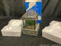 Miniature decorative greenhouse water feature with glass sides and stone-patterned base, shown with original foam packaging inserts and blue box.