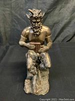 Front view of seated horned devil statue playing a pan flute, showing detailed beard, horns, and fur-covered legs on rock base.