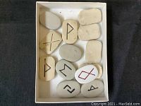 Box containing 14 light-colored stones with various black and red engraved symbols resembling runes.