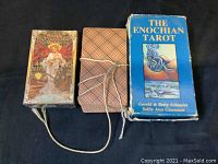 Photo showing three tarot card decks: Golden Art Nouveau sealed deck, unknown deck wrapped with string with plaid back cards, and The Enochian Tarot deck in a blue box.