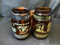 Photo showing two brown glass mugs with wooden handles side by side. Mugs have metallic bands and distinct scenic prints: Wildwood-by-the-Sea and St. Johns Church Elora.