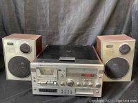 Front view of the Yorx stereo receiver with two matching speakers on either side, showing the control panel, cassette slot, analog tuning dial, digital display, and speaker grilles.