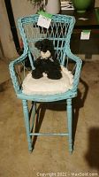 Front view of light blue antique child's chair with white cushion and black plush toy dog placed on seat. Chair is rustic with woven details.