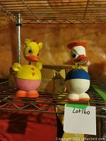 Donald Duck and Daisy Duck antique paper mache candy containers on a metal shelf with cardboard boxes in the background and orange-brown wall.