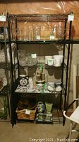 Full front view of black metal baker's rack with multiple shelves and various items on each shelf. Walls and floor background indicate basement location.