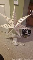 White Christmas star lamp on white metal stand with electrical cord and switch, viewed from front.