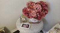 White wicker round table holding a pink hydrangea floral arrangement, small silver clock and framed floral picture