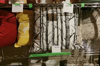 Five decor pillows arranged on metal shelving, showing red, yellow, black and white birch tree pattern, and textured beige pillow.